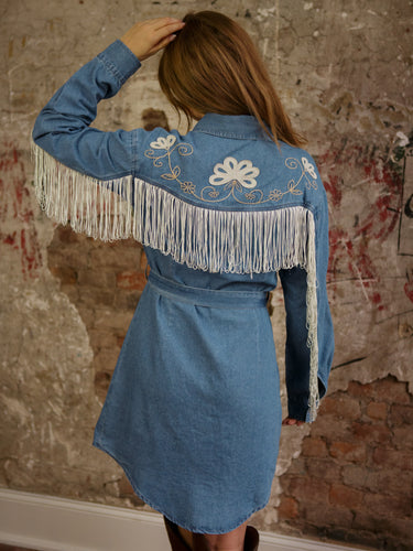 Denim mini dress with long sleeves, pearl snap buttons, floral embroidery, fringe details, and waist belt.