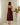 A woman is shown from behind wearing a burgundy satin slip dress with lace detailing at the back, standing in front of white curtains next to a decorative floral arrangement on a white pedestal.
