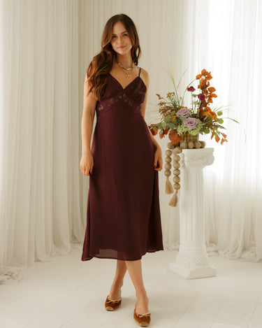 A woman modeling a sleeveless burgundy midi dress with lace detailing on the bodice, standing in a bright room with white curtains and a decorative floral arrangement on a pedestal.