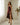 A woman wearing a dark burgundy satin slip dress with a thigh-high slit and lace detail at the neckline, standing in a bright room with white curtains and a floral arrangement.