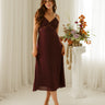 A woman modeling a sleeveless burgundy midi dress with lace detailing on the bodice, standing in a bright room with white curtains and a decorative floral arrangement on a pedestal.