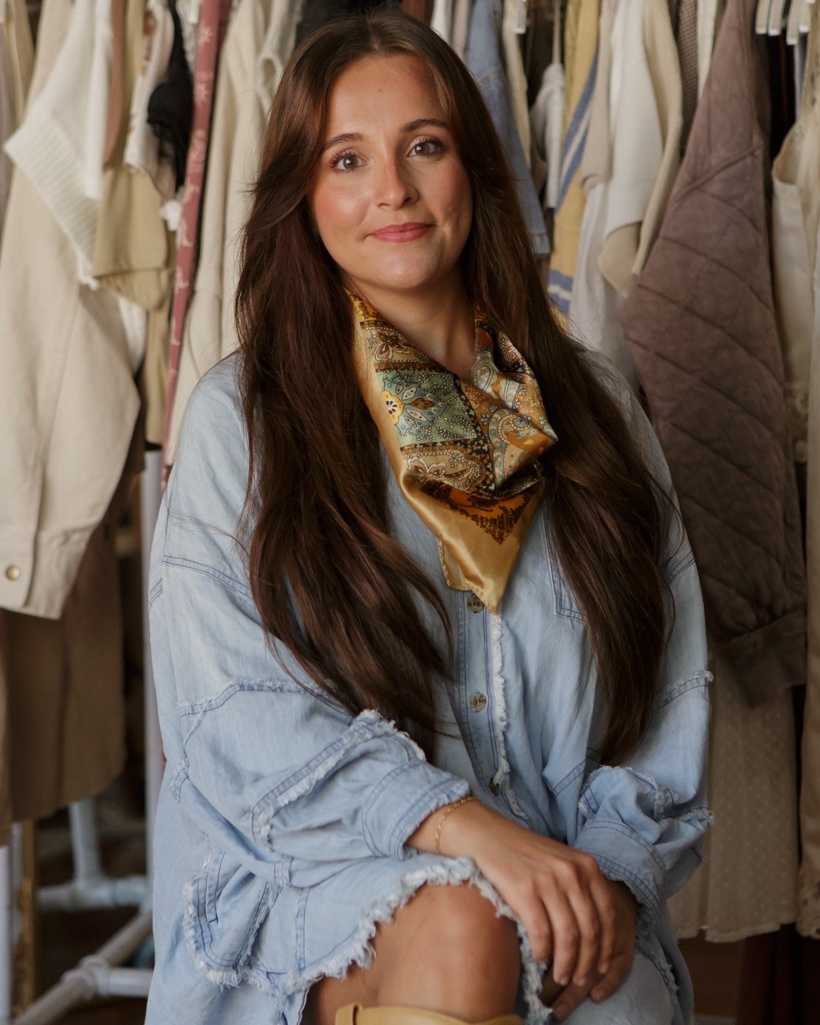 Alyssa Messina in an oversized denim dress with a paisley silk scarf, in front of a clothing rack